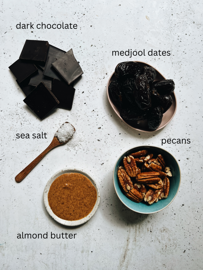 ingredients on a table. Dark chocolate pieces in a pile, medjool dates on a plate, a teaspoon of sea salt, a bowl of pecans and a bowl of almond butter.