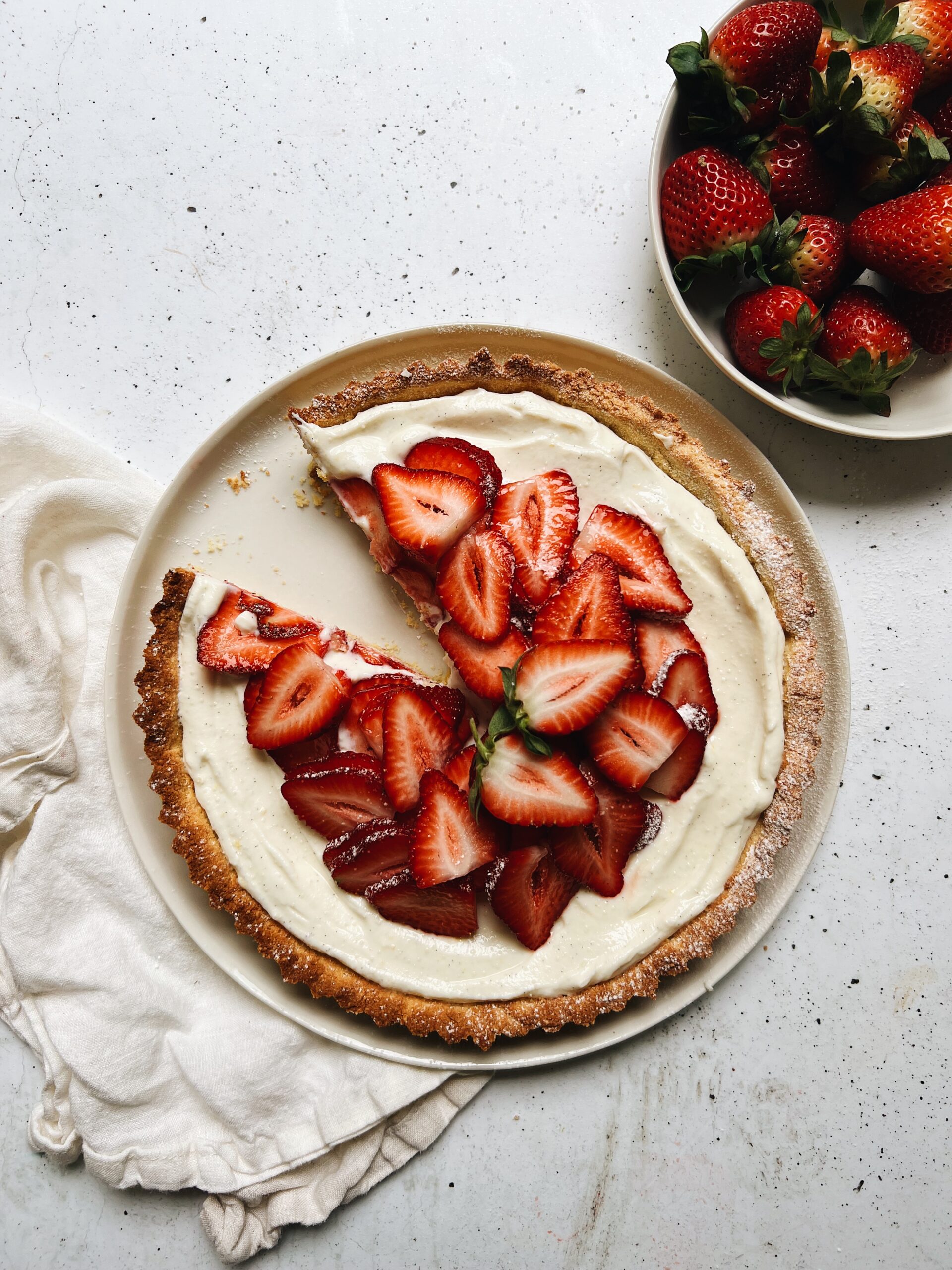 Strawberry tart sits on napkin on table. A bowl of strawberries sit alongside.