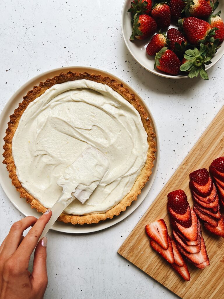 Hand filling ricotta into tart base. Chopped strawberries sit on a chopping board alongside. 
