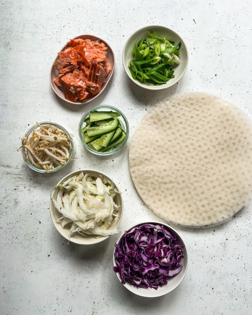 Rice paper sheets surrounded by small bowls of salmon, onion, sprouts, purple cabbage and cucumber