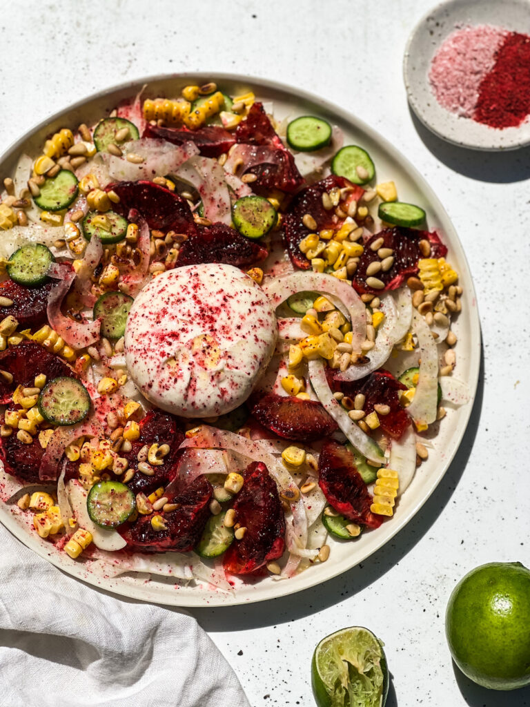 Burrata With Blood Orange, Fennel, Corn And Ooray Powder on a plate surrounded by limes