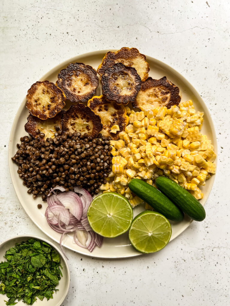 A plate filled with lentils, corn, grilled squash, lime halves, raw red onion and baby cucumbers.
