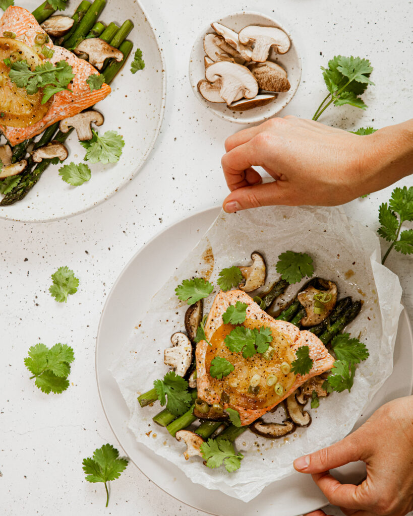 Hands open the bake paper parcel, with salmon, mushroom and asparagus. 